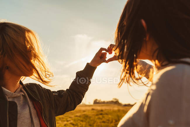Visão lateral de jovens amigas olhando umas para as outras e fazendo gesto cardíaco com as mãos contra o pôr do sol na natureza — Fotografia de Stock