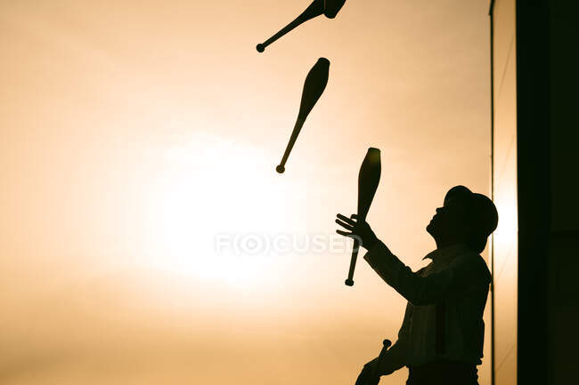 Low angle side view of unrecognizable male circus artist in hat juggling clubs against sunset sky — Stock Photo