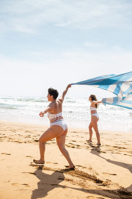 Visão lateral de corpo inteiro de amigas em trajes de banho passeando na costa arenosa com toalha perto do oceano sob o céu azul nublado em dia ensolarado — Fotografia de Stock