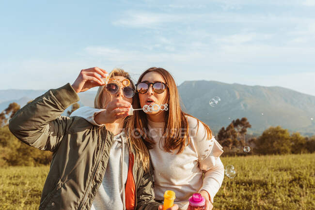 Fermez vos amies dans des lunettes de soleil soufflant des bulles de savon ensemble debout dans l'étreinte sur la prairie dans les montagnes — Photo de stock