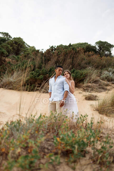 Вид сбоку любящей многорасовой пары молодоженов, обнимающихся, стоя на песчаном холме в день свадьбы — стоковое фото