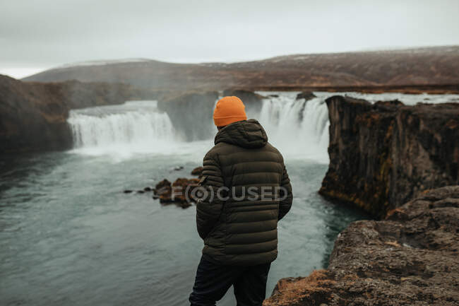 Back view of young tourist in winter wear looking at cascade and mountain river between stone hill — Stock Photo