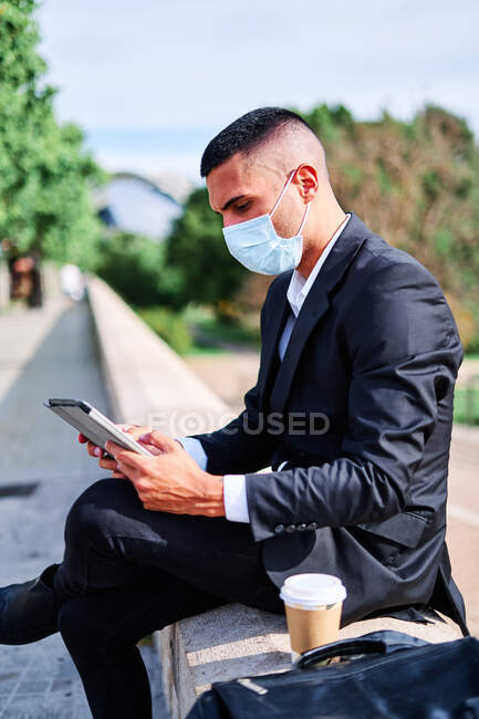 Вид збоку концентрованого молодого бізнесмена в елегантному костюмі і захисній масці читання новин на планшеті, маючи перерву з чашкою кави на міській вулиці під час пандемії коронавірусу — стокове фото