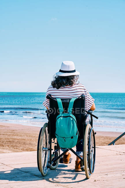 Visão traseira de viajante feminino irreconhecível em cadeira de rodas com mochila desfrutando de viagem de verão na praia perto do mar azul — Fotografia de Stock