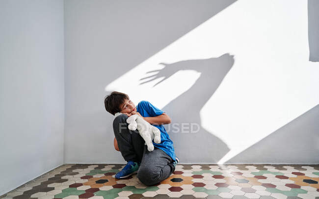 Niño asustado con juguete en las manos sentado cerca de la pared con sombra de padre enojado violento con el brazo levantado - foto de stock
