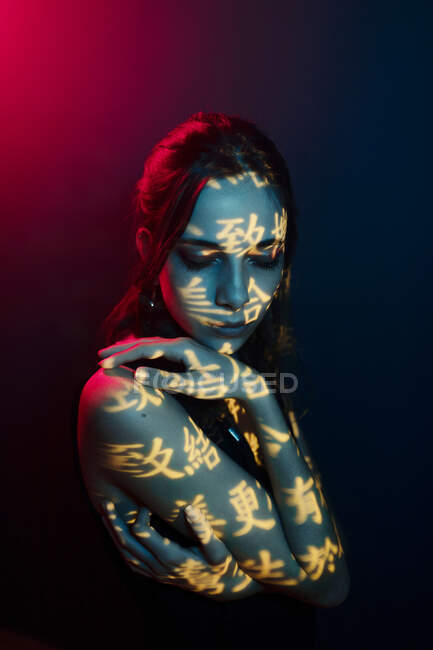 Fashionable young female model with light projection in shape of oriental hieroglyphs looking down in dark studio with red illumination — Stock Photo
