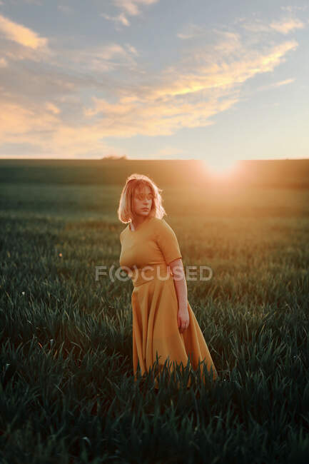 Joven hembra en vestido vintage mirando hacia otro lado pensativamente mientras está de pie solo en el campo de hierba al atardecer en la noche de verano en el campo - foto de stock