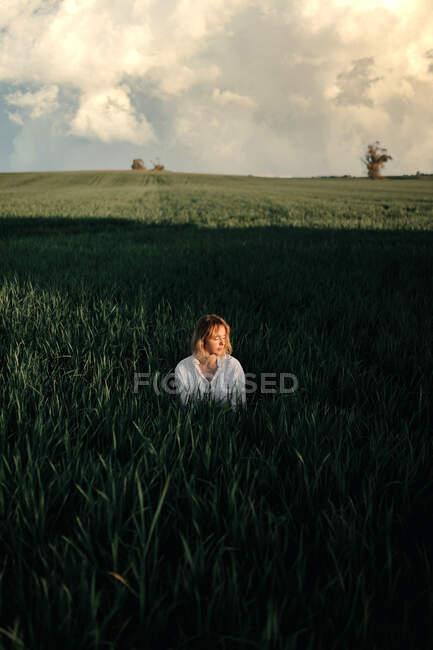 Peaceful young female in retro styled white blouse sitting amidst tall green grass and closed eyes while resting in summer evening in countryside — Stock Photo
