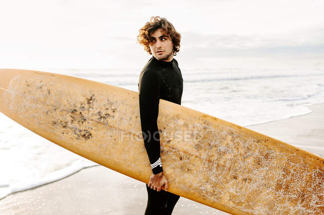 Vista lateral del hombre surfista vestido con traje de neopreno de pie mirando hacia otro lado con la tabla de surf en la playa durante el amanecer en el fondo - foto de stock