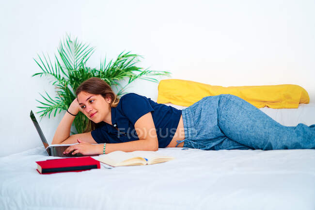 Jeune pigiste positive en vêtements décontractés couchée sur le lit et tapant sur un ordinateur portable tout en travaillant sur un projet à distance à la maison — Photo de stock