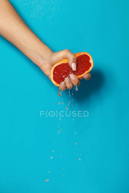 Crop anonymous female with manicure squeezing half of fresh ripe juicy grapefruit against blue background — Stock Photo