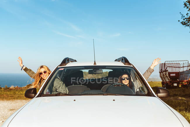 Cheerful females sitting in car and waving hands out of windows having road trip on shore of sea — Stock Photo