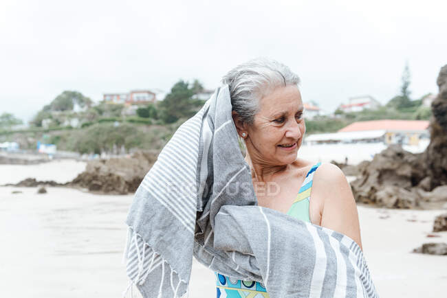 Hembra envejecida positiva en traje de baño limpiando el cabello mojado con toalla después de nadar en el mar en el día de verano - foto de stock