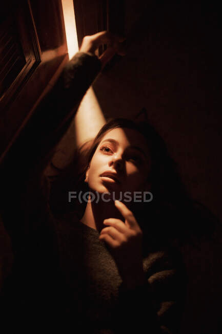 Dall'alto di silenziosa giovane femmina sdraiata sul pavimento nella stanza buia con la luce che brilla dalla porta aperta guardando altrove — Foto stock