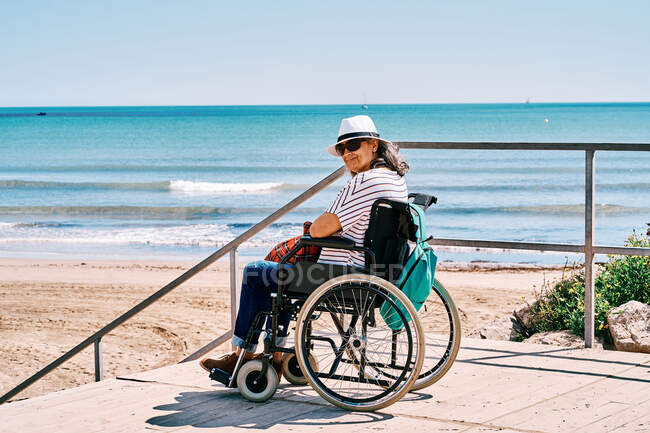 Seitenansicht Reisenden im Rollstuhl mit Rucksack genießen Sommerreise am Strand in der Nähe des blauen Meeres Blick auf Kamera — Stockfoto