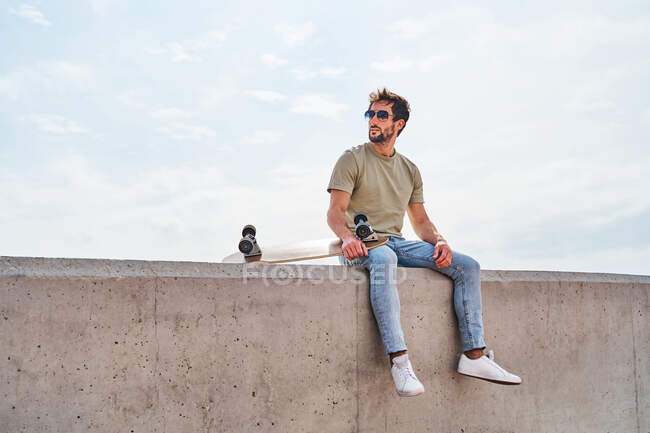 Ganzkörper ernsthafter bärtiger Mann in lässiger Kleidung und Sonnenbrille sitzt mit Skateboard auf Betonkante gegen plätscherndes Meer — Stockfoto