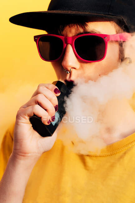 Femme contemporaine dans une tenue élégante expirer les fumées tout en se tenant près du mur jaune et vaping sur la rue de la ville — Photo de stock