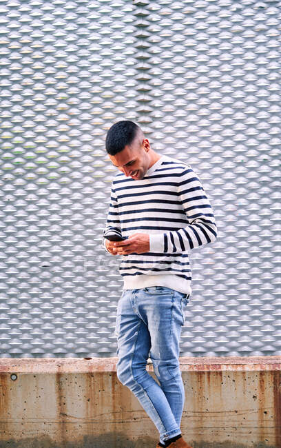 Hombre hispano con elegante atuendo mirando hacia otro lado y usando el teléfono celular mientras se apoya en la pared en la calle de la ciudad - foto de stock