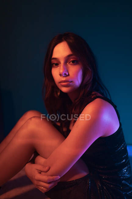 Seitenansicht des ruhigen jungen weiblichen Modells in Kleid sitzt auf dem Boden und schaut in die Kamera im dunklen Studio mit bunten Lichtern — Stockfoto