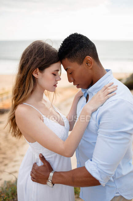 Вид сбоку любящей многорасовой пары молодоженов, обнимающихся, стоя на песчаном холме на фоне моря в день свадьбы — стоковое фото
