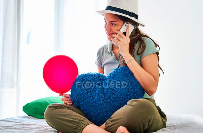 Молода жінка в повсякденному одязі і капелюсі тримає червону кульку і серцеподібну подушку, переглядаючи на смартфоні, святкуючи день народження наодинці на ліжку з їжею і соком — стокове фото