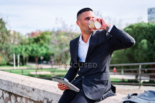 Vista lateral do jovem homem hispânico feliz em terno formal elegante beber bebida takeaway e tablet de navegação, tendo tempo livre e descansando no parque no dia ensolarado de verão — Fotografia de Stock
