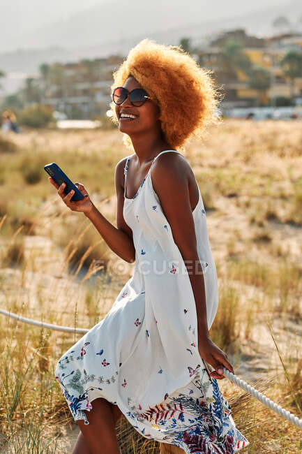 Бічний вид веселої афроамериканської жінки з африканською зачіскою в сонцезахисних окулярах і літнє вбрання, що дивиться на смартфон на пляжі. — стокове фото