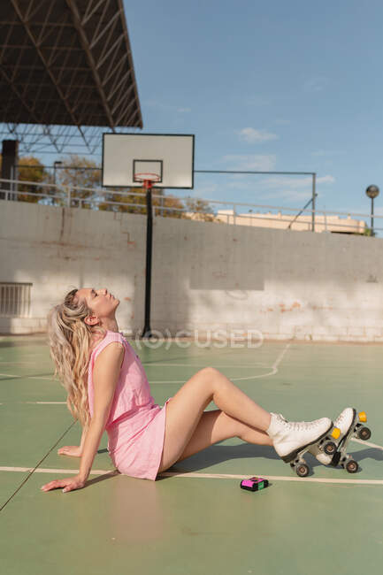 Vista lateral de longitud completa de la hembra en forma de vestido de rosa y patines de ruedas sentado con los ojos cerrados en el campo de deportes soleado - foto de stock