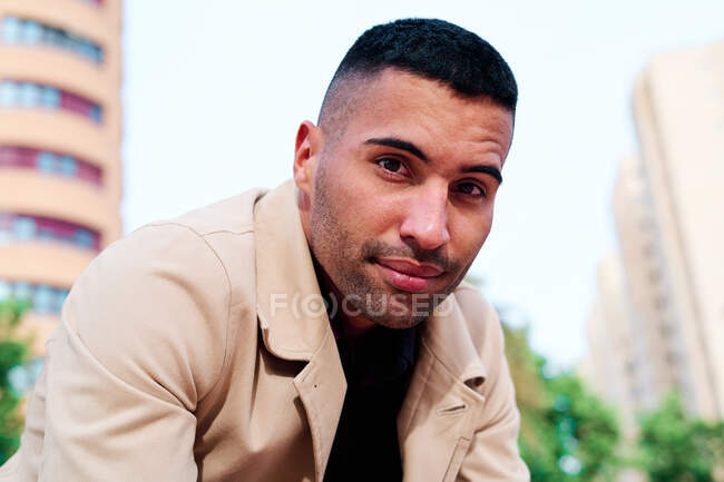 Handsome young unshaven Hispanic male in stylish jacket looking at camera while sitting on modern city street — Stock Photo