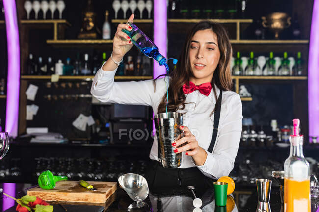 Camarera femenina enfocada en traje elegante añadiendo líquido de la botella en jigger mientras prepara cóctel de pie en el mostrador en el bar moderno - foto de stock