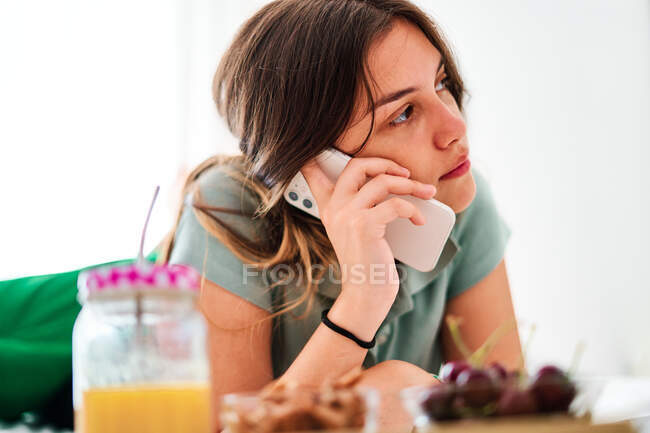Vue latérale de la jeune étudiante parlant sur un téléphone portable près de la table avec des fruits frais et du jus tout en passant la matinée à la maison — Photo de stock
