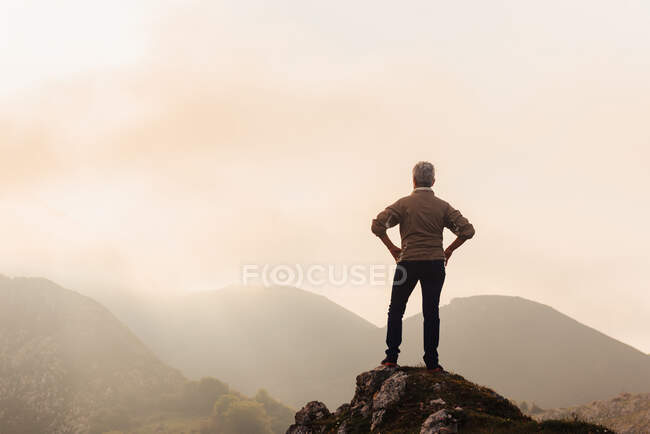 Вид ззаду анонімного дослідника з руками на талії, захоплююча гірська місцевість на тлі хмарного східного неба вранці в природі — стокове фото
