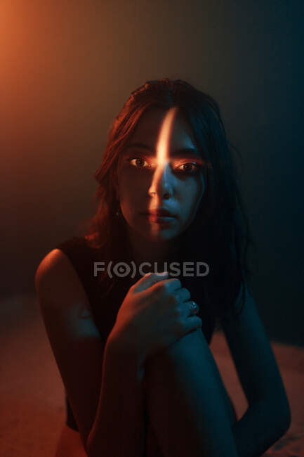 Junges, emotionsloses weibliches Modell mit kreuzförmiger Lichtprojektion auf das Gesicht, das im dunklen Studio sitzt und in die Kamera blickt — Stockfoto