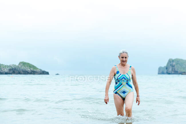 Positive active fit aged female in swimsuit walking out of sea water while enjoying summer day on beach — Stock Photo