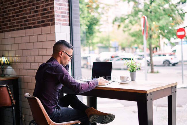 Gerente masculino hispano serio en camisa violeta y gafas escribiendo en el teclado netbook mientras está sentado en la mesa detrás de la ventana y trabajando en el proyecto en la cafetería - foto de stock
