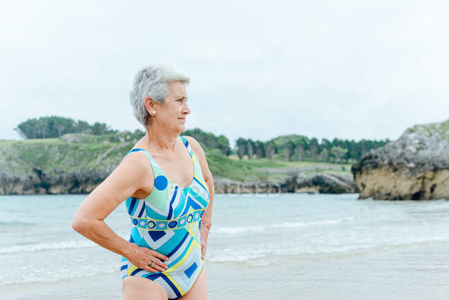 Ältere Frau mit durchtrainiertem Körper trägt einen eleganten, farbenfrohen Badeanzug mit geometrischem Print und steht mit den Händen an der Taille an einem Sommertag gegen das Meer — Stockfoto