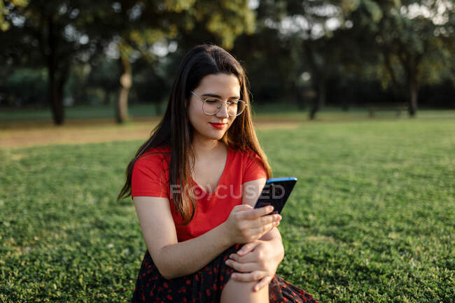 Femme en vêtements d'été assis sur la prairie verte dans le parc et surfer sur Internet sur téléphone mobile tout en divertissant le week-end dans la soirée — Photo de stock