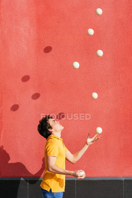 Vista lateral de un joven talentoso macho realizando truco con bolas malabares mientras está de pie en el pavimento cerca de la pared roja brillante - foto de stock
