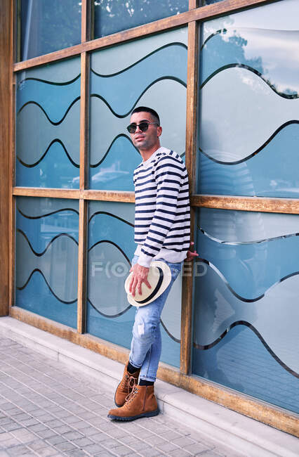 Full body adult Hispanic male in trendy outfit leaning on glass wall with abstract ornament and looking away — Stock Photo