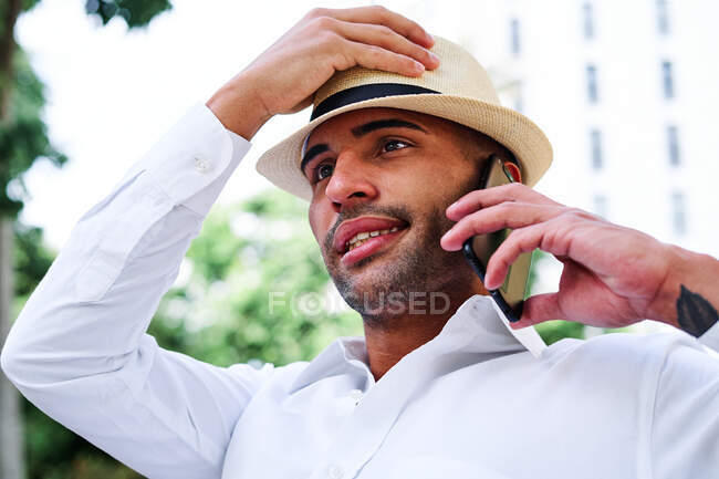 Низький кут усмішки себе запевнив молодого бородатого іспанського джентльмена в класичному одязі і капелюх, який розмовляє по мобільному телефону, відпочиваючи на лавці в місті. — стокове фото