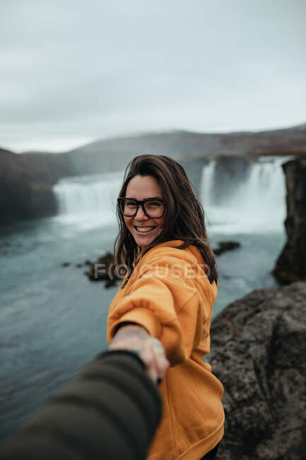 Вид сбоку юного жизнерадостного туриста в очках с пением, держащего руку человека и смотрящего в камеру на холме возле водопада и горной реки — стоковое фото