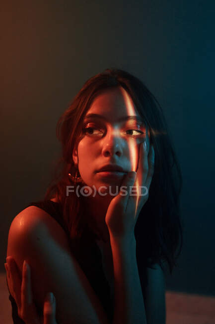 Joven modelo femenino pensativo con proyección de luz en la cara sentado en estudio oscuro y mirando hacia otro lado - foto de stock