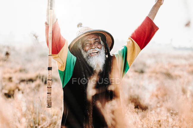 Cheerful old ethnic rastafari with dreadlocks with eyes closed celebrating victory while standing in a dry meadow in the nature — Stock Photo