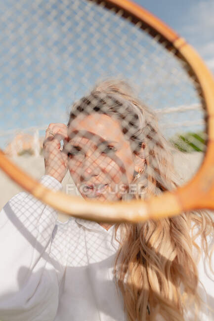 Позитивна молода жінка в білому одязі торкається волосся і дивиться на камеру через тенісну ракетку, стоячи на сонячній вулиці — стокове фото