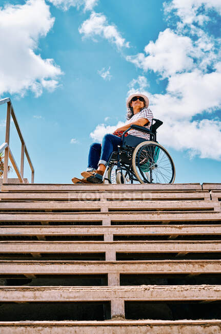 Bas angle corps complet de femme handicapée positive assis dans un fauteuil roulant près de l'escalier regardant loin contre le ciel bleu en ville — Photo de stock