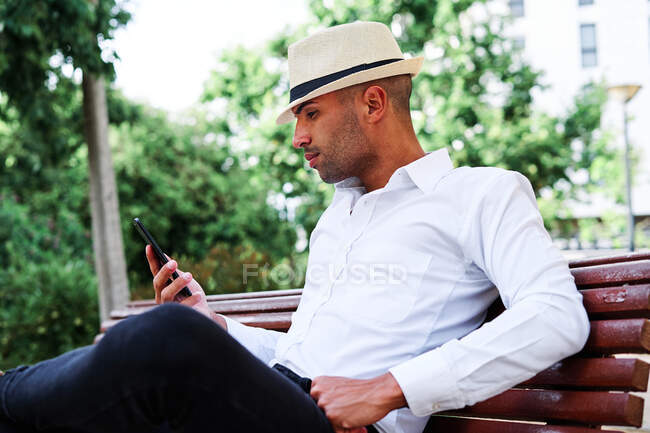 Low angle of smiling self assured young bearded Hispanic gentleman in classy outfit and hat texting on mobile phone while resting on bench in city — Stock Photo