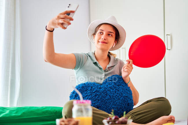 Full length of young female in casual clothes and hat holding red balloon and heart shaped cushion and taking selfie on smartphone while celebrating birthday alone on bed with food and juice — Stock Photo
