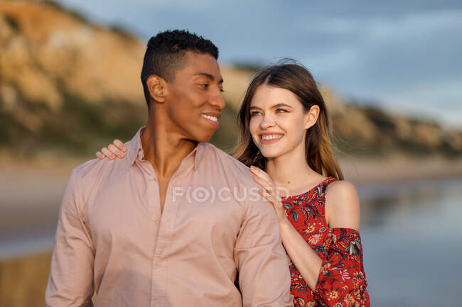Loving woman hugging black man from behind while spending summer day together on seashore — Stock Photo