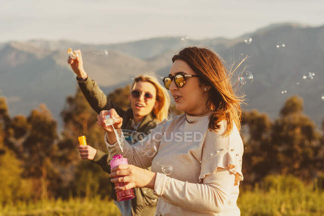 Близкие подруги в солнечных очках, дующие мыльные пузыри вместе стоя на лугу в горах — стоковое фото