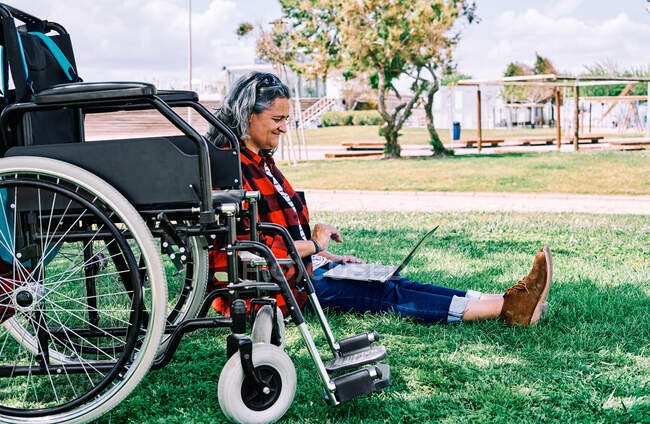 Повне тіло позитивної сіроволосистої жінки з ноутбуком на колінах, що сидить на зеленій траві біля інвалідного візка в парку — стокове фото
